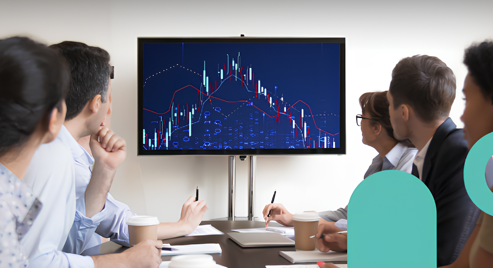 Five business professionals seated around a conference table attentively looking at a large monitor displaying a complex financial chart with red and white trends on a dark blue background, while some hold pens and coffee cups.