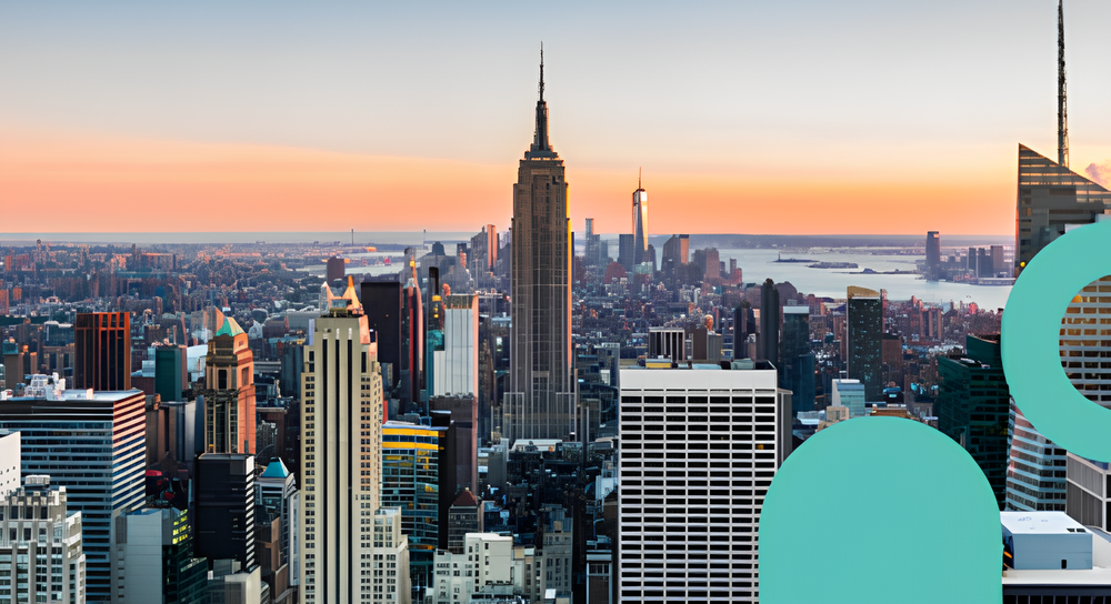 Panoramic view of New York City skyline at sunset with the Empire State Building prominently in the center and One World Trade Center visible in the background near the water.