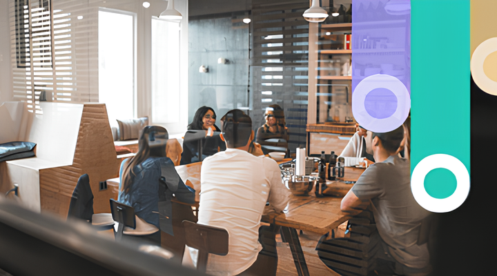 Group of six diverse people sitting around a wooden table in a modern office space, engaged in a discussion during a meeting.