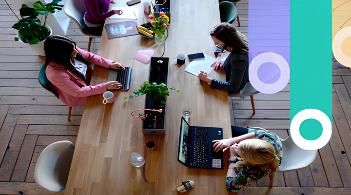 Overhead view of four people working collaboratively at a wooden table with laptops, notebooks, and coffee cups, in a bright, modern office space with wooden floors and plants.