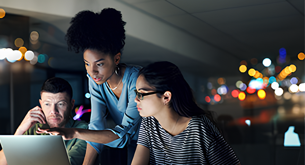 Three diverse colleagues working together at a laptop in a modern office at night, with city lights blurred in the background.