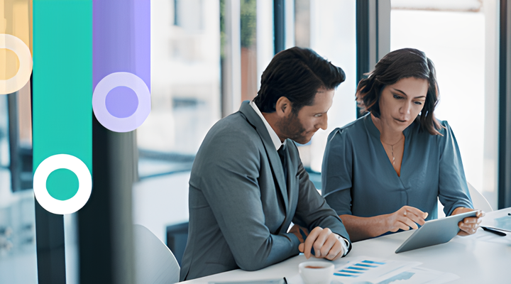 Two business professionals sit at a table by large windows, reviewing data on a tablet with printed charts in a bright office.