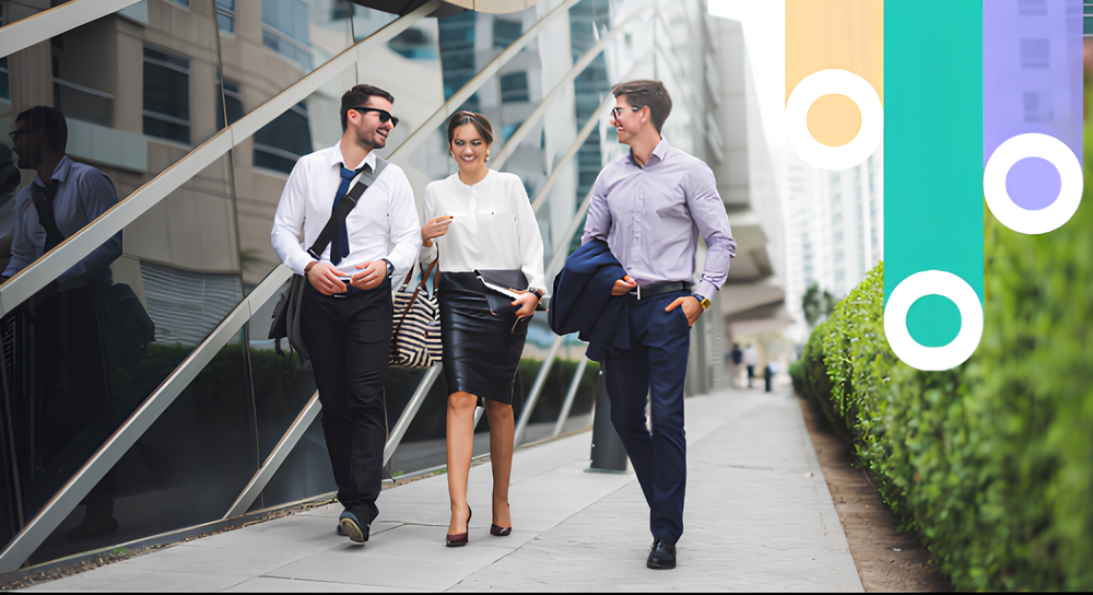 Three young business professionals walking together on a city sidewalk next to a modern glass building, smiling and engaged in conversation, two men on either side of a woman, all dressed in business attire.