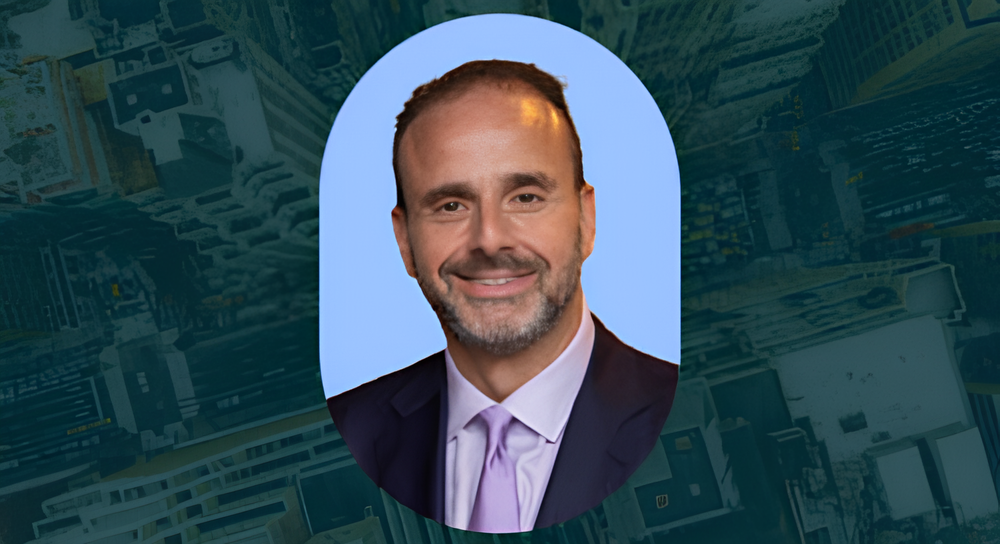 Smiling man with short dark hair and beard wearing a dark suit, white shirt, and purple tie against a light blue oval background with a dark, abstract cityscape behind.