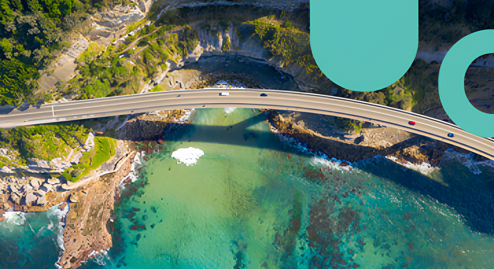 Aerial view of a curved bridge crossing clear turquoise water with rocky coastline and green vegetation on the shore.