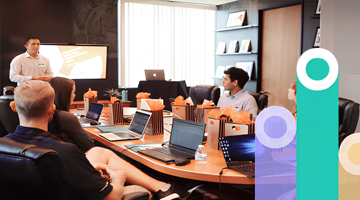 A business meeting in a conference room with five people seated around a wooden table with laptops and gift bags, listening to a man standing and presenting information displayed on a screen.