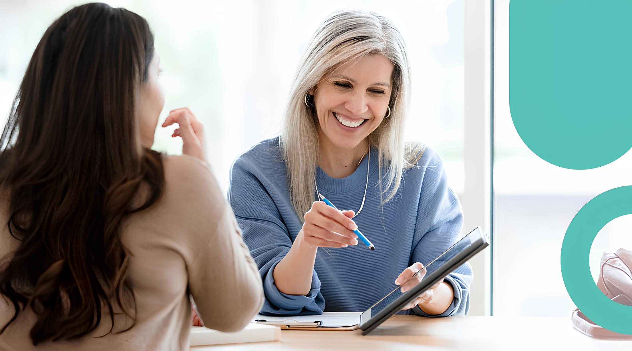 Smiling woman with blonde hair and blue sweater holding a tablet and pointing at the screen with a stylus while talking to another woman with long brown hair wearing a beige sweater, seated at a table near a window.