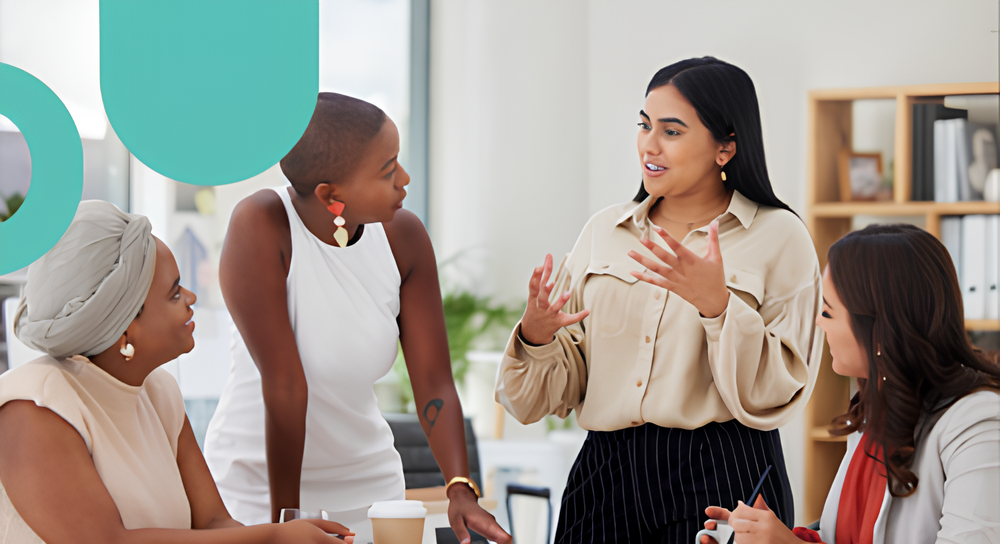 Four diverse women engaged in a discussion in a modern office setting, with one woman standing and gesturing while the others listen attentively.