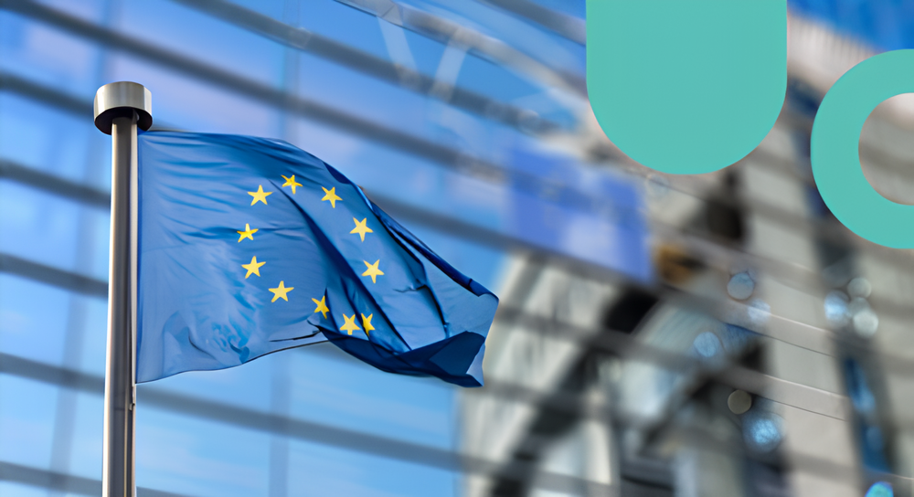 European Union flag with yellow stars on a blue background, waving on a flagpole in front of a modern reflective glass building.