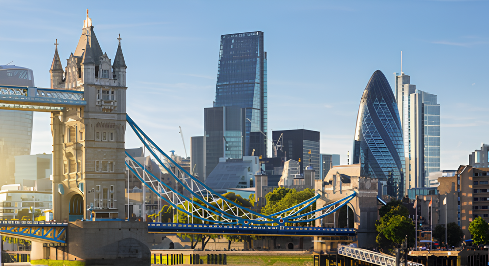 View of London's Tower Bridge with its twin towers and blue suspension cables against a clear sky, with modern skyscrapers including the Gherkin and the Leadenhall Building in the background.