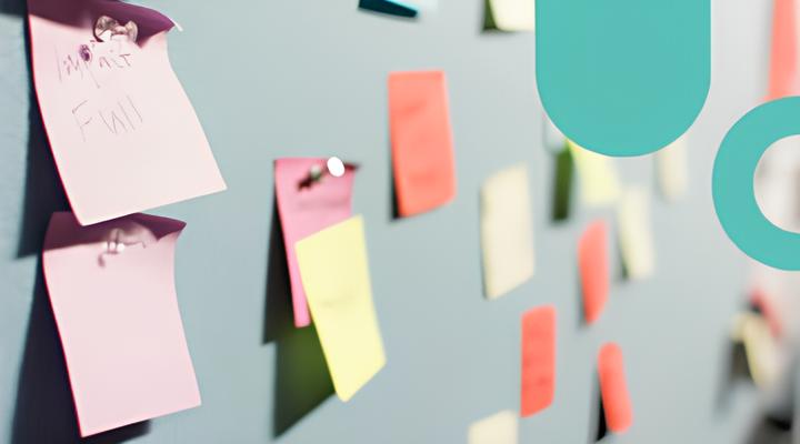 A light gray wall with various colorful sticky notes pinned on it, including pink, yellow, orange, and green notes, some overlapping and out of focus, with one pink note in the foreground showing partially readable handwriting.