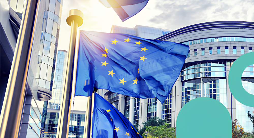 European Union flags waving in front of modern glass buildings with sunlight shining through.