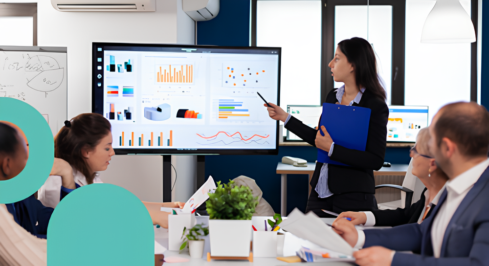 A businesswoman in a black suit holding a blue clipboard and pen points at a large screen displaying various colorful charts and graphs during a meeting with four colleagues seated around a table.