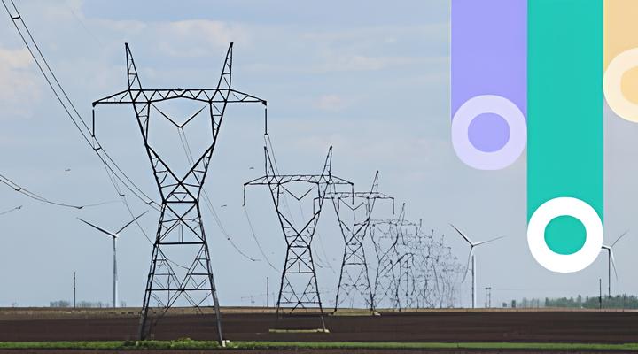 A series of tall electrical transmission towers with cables stretching across a flat field under a partly cloudy sky, with wind turbines visible in the distance.