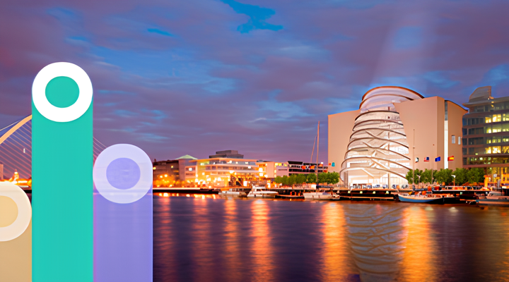Illuminated modern buildings and boats reflected on calm water at dusk under a cloudy sky.