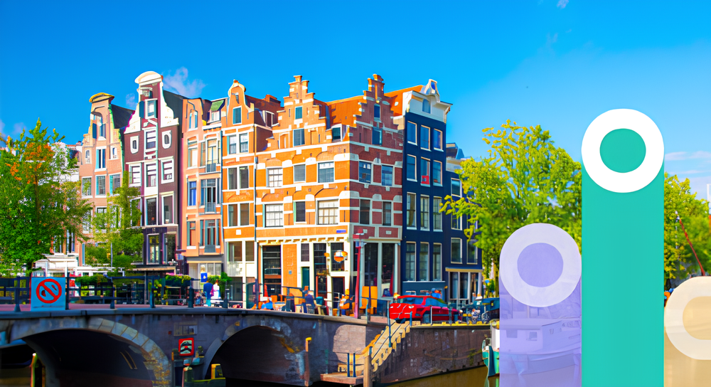 Colorful traditional narrow buildings with stepped gables along a canal in Amsterdam, with a stone bridge crossing the water and green trees under a blue sky.