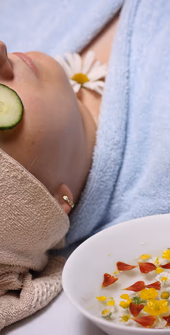 Person lying down with a cucumber slice on one eye, wearing a beige head wrap and surrounded by a blue towel, next to a bowl of water with floating flower petals.