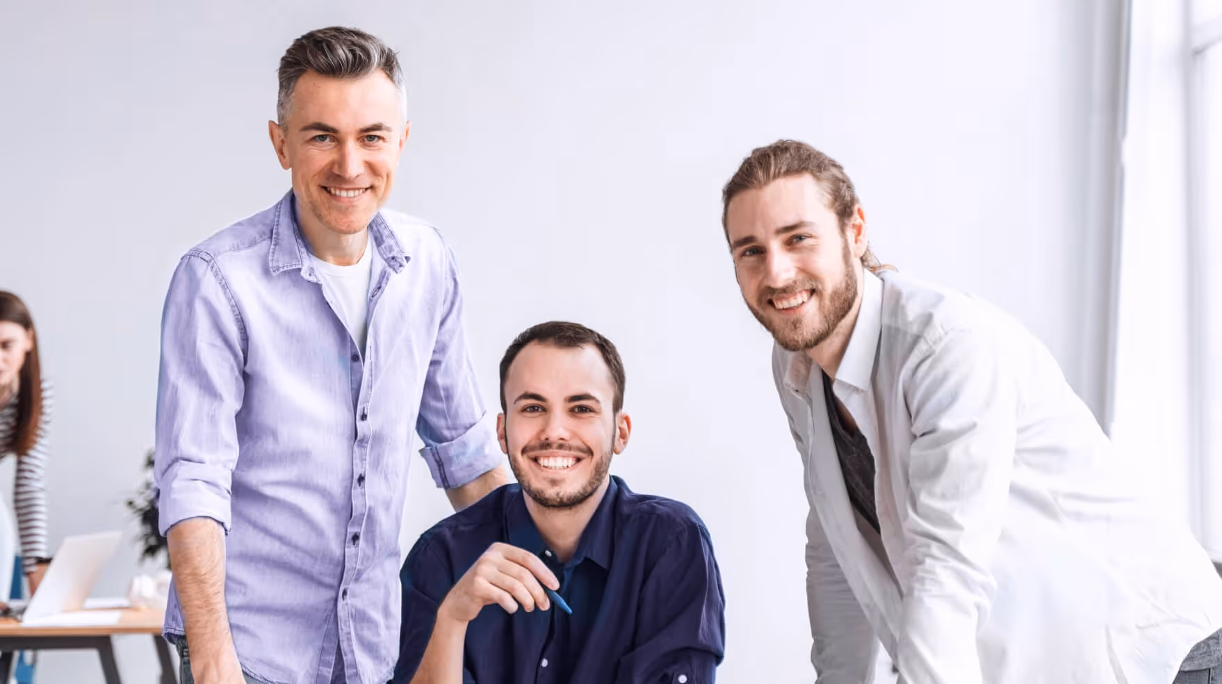 Three smiling men posing together in a bright office, two standing and one seated holding a pen.