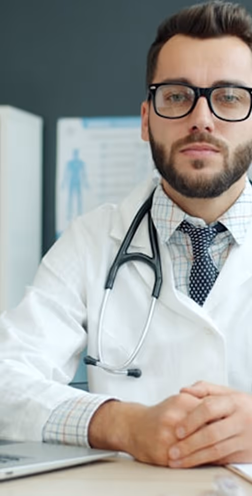 Male doctor wearing glasses and a white coat with a stethoscope around his neck, sitting at a desk with hands clasped.