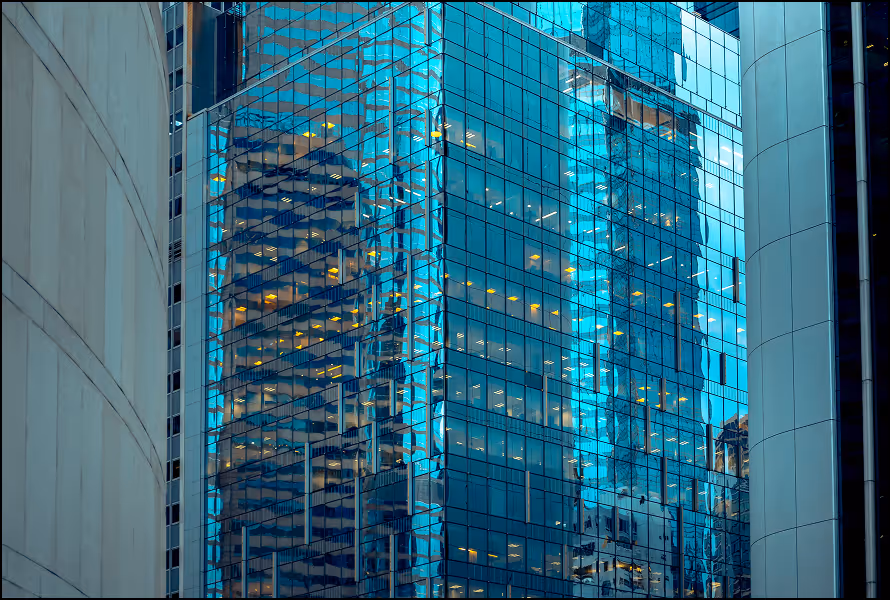 Close-up of modern glass office building reflecting another skyscraper and surrounding structures.