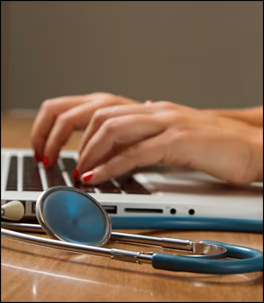 Hands with red nail polish typing on a laptop keyboard with a stethoscope resting on the wooden table.