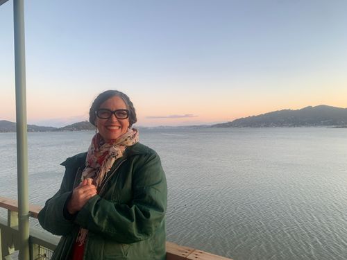 Alison Alstrom, a white woman with glasses and brown hair, standing in front of a large body of water. She is wearing a scarf and a green jacket.