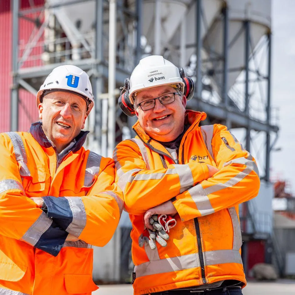 Een foto van een vrouw en een man op een werkplaats met grote gele steigers. De vrouw wijst iets aan. Daarover staat het logo en een quote van BAM Infra