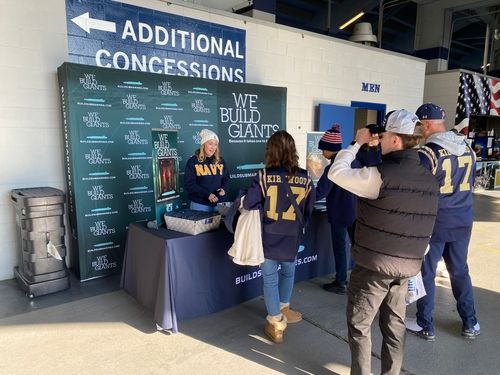 A woman in a Navy shirt stand behind a table and in front of a backdrop with BuildSubmarines.com on it. Fans approach the table.