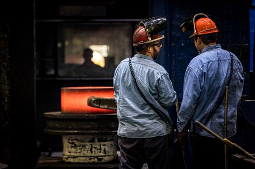 Two men stand facing a hot piece of manufacturing machinery. They are wearing helmets with face shields, but the shields are up while they talk.