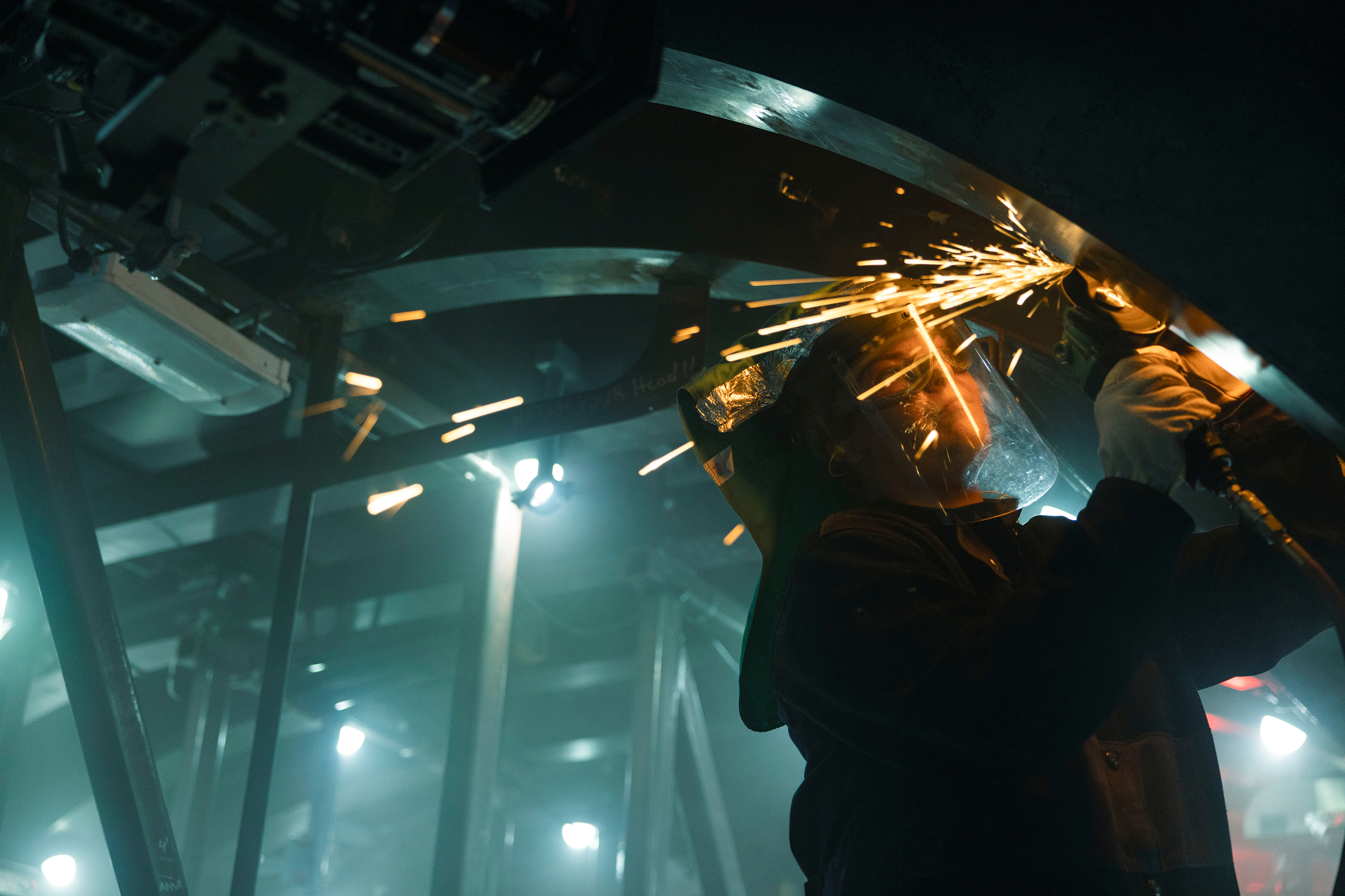 A woman wearing a blue shirt lifts the shield of the welding helmet she's wearing to reveal her face.