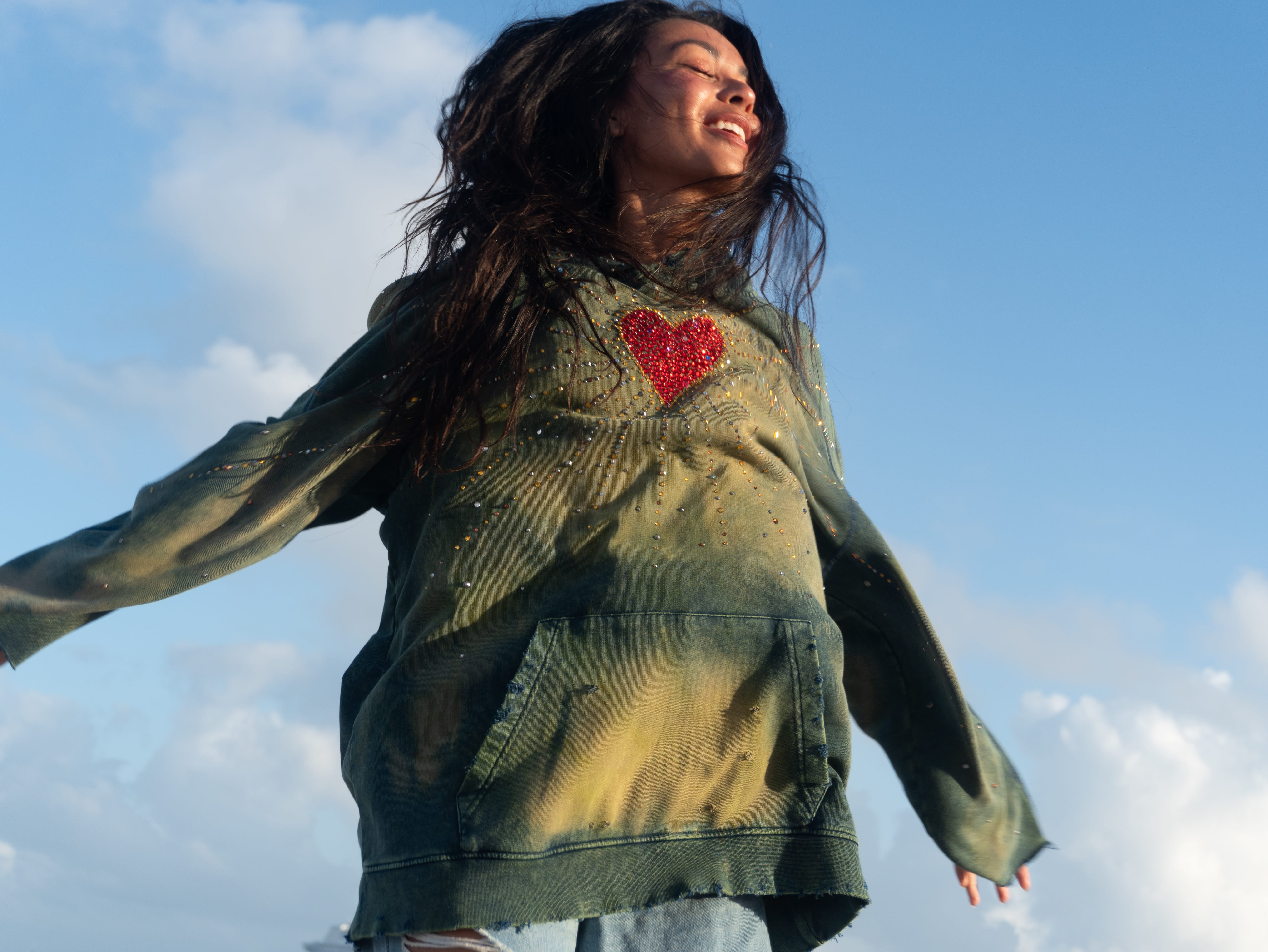 Woman wearing a green hoodie with a red heart design, sitting on a concrete ledge by the sea.