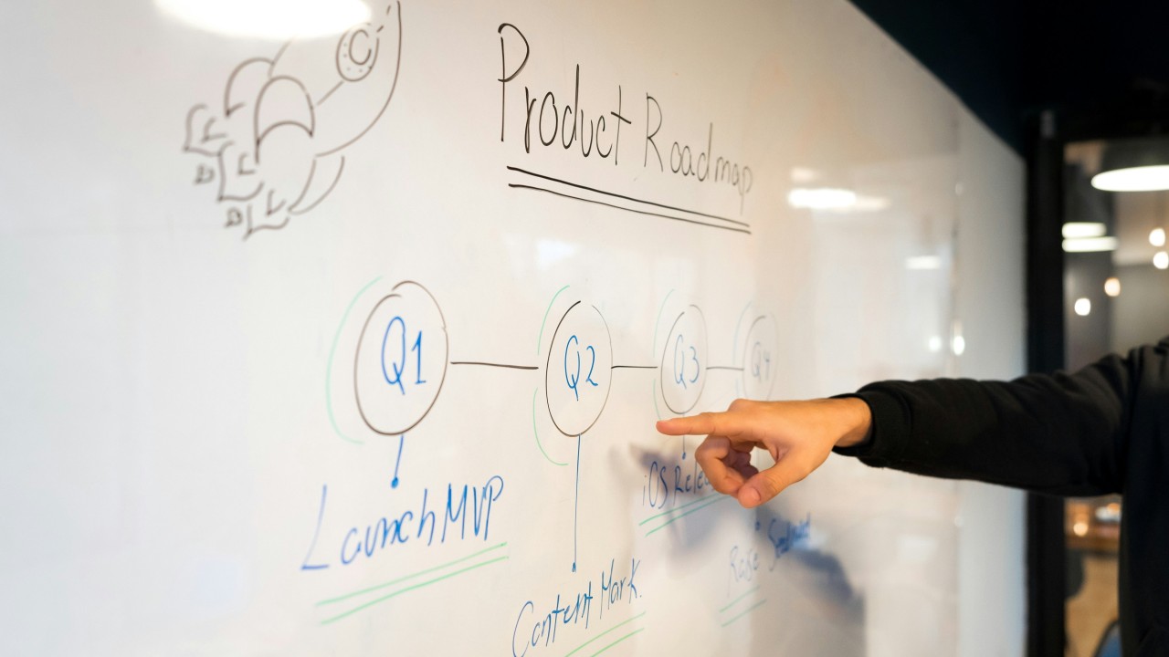 Graphic featuring a person pointing at a product roadmap on a whiteboard, overlaid with the bold title: “THE 1% QUESTION: WHEN VCs ACTUALLY EARN THEIR SEAT AT THE TABLE.” Orange horizontal lines appear along the bottom.