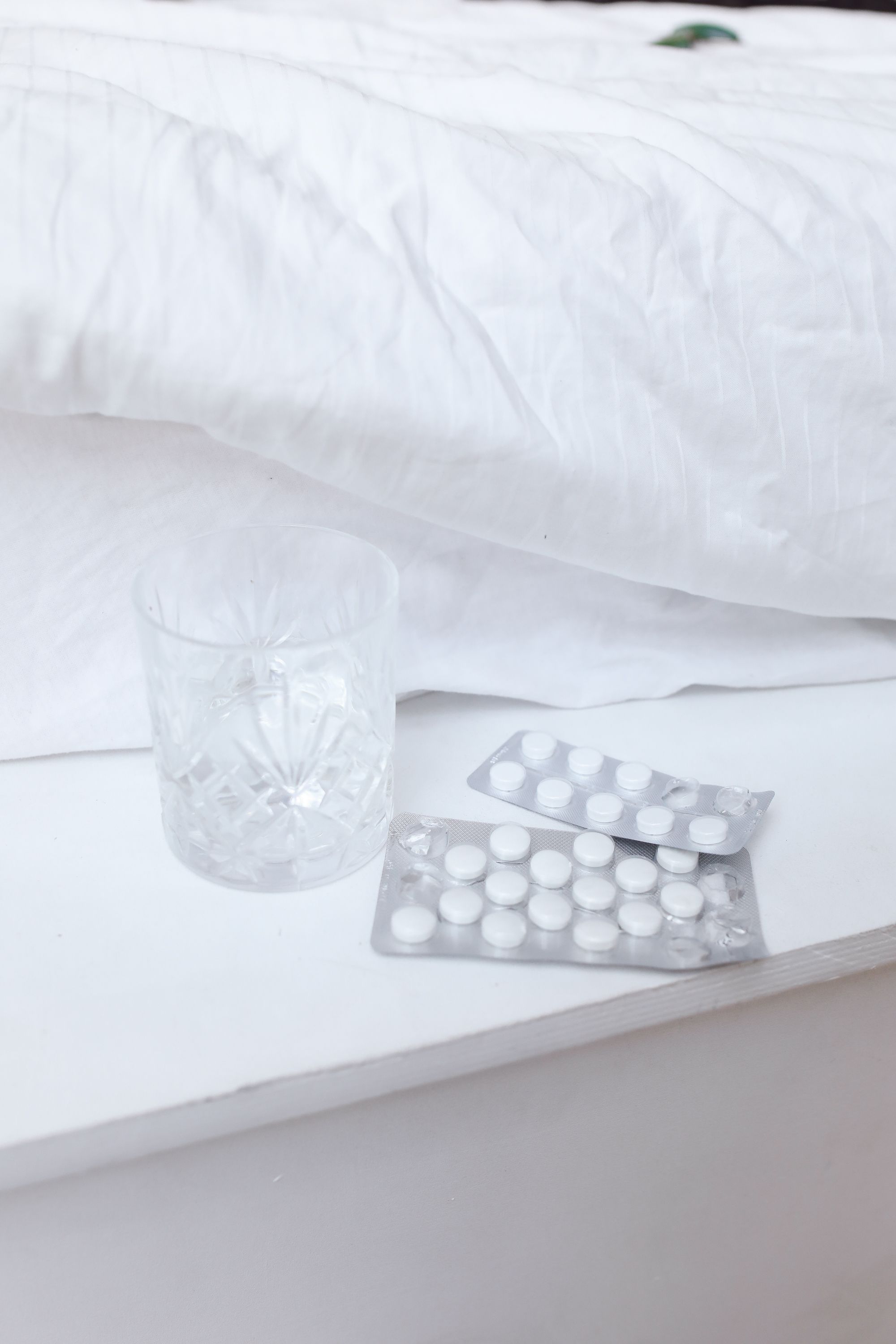 A clear glass and tablet blister packs on a white nightstand by a bed. Photo by Polina Tankilevitch from Pexels.