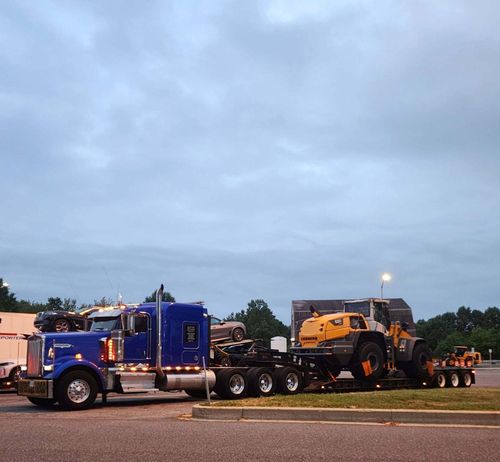 Blue Kenworth heavy-haul truck from RCS Trucking transporting a large yellow Liebherr loader on a flatbed trailer at dusk, showcasing the company’s owner-operator fleet capabilities.