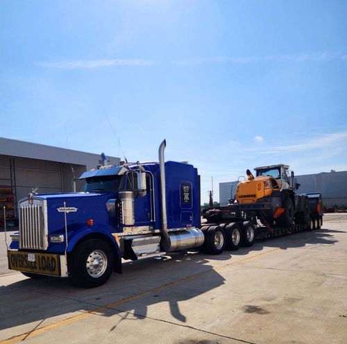RCS Trucking blue Kenworth flatbed truck hauling oversized construction equipment in daylight at company terminal, representing owner-operator opportunities with RCS Trucking in Virginia.
