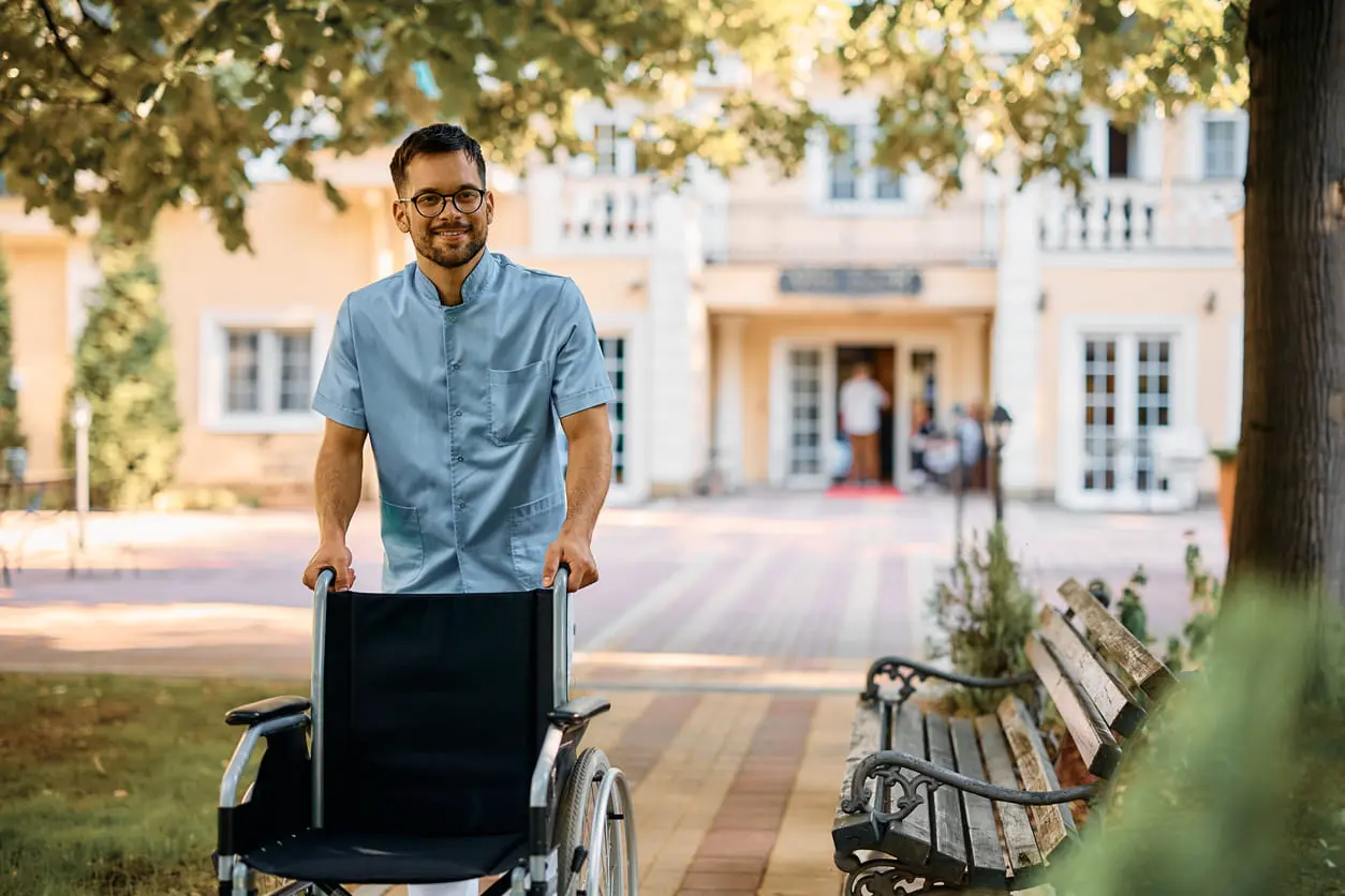 Glückliche männliche Krankenschwester, die den Rollstuhl durch den Park eines Pflegeheims schiebt