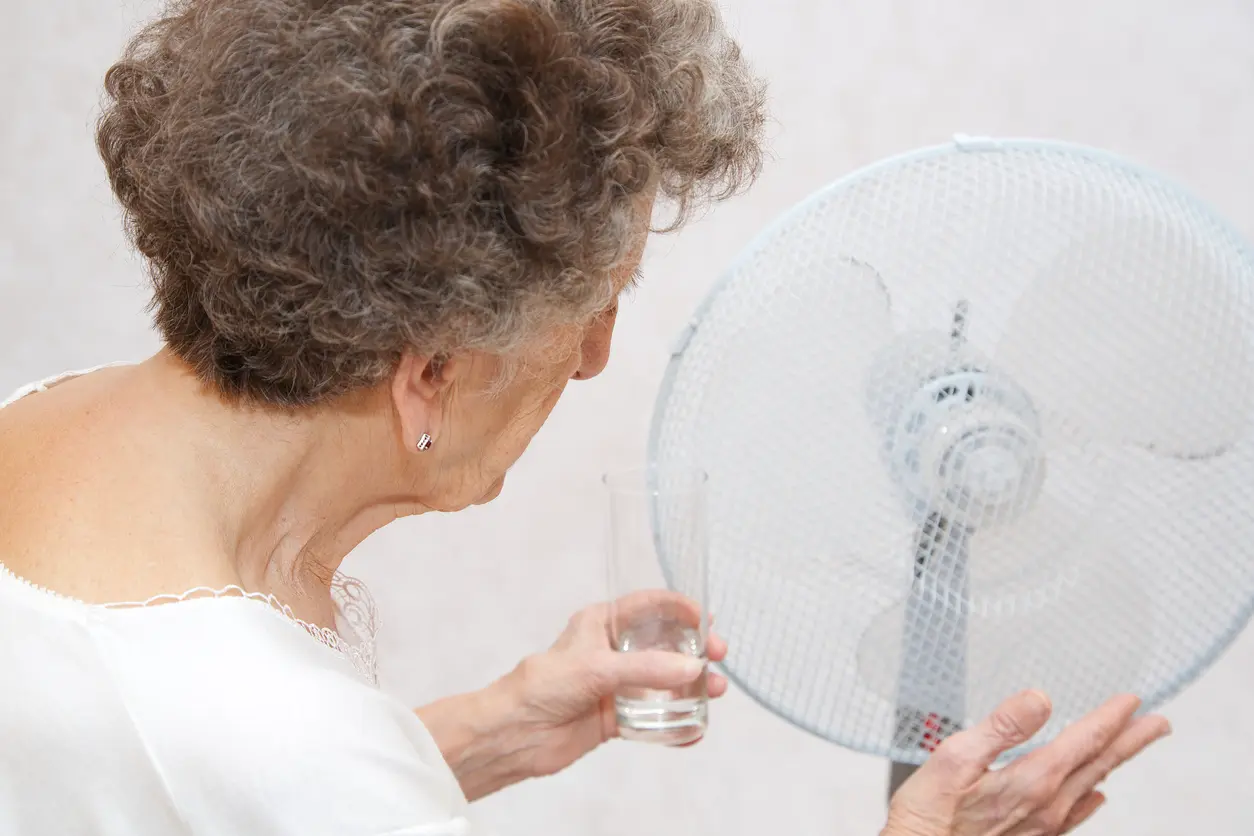 Ältere Frau kühlt sich am Ventilator ab mit Wasserglas