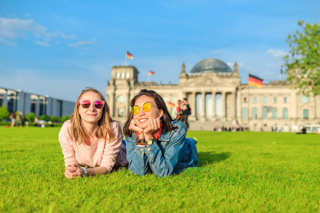 Zwei junge glückliche Mädchen, die eine Sonnenbrille tragen, die auf einem Gras liegt und vor dem Bundestagsgebäude in Berlin ihren Spaß haben.