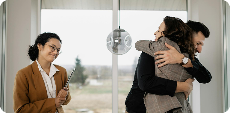 A couple embracing in a bright office setting while a professional stands nearby holding a clipboard.