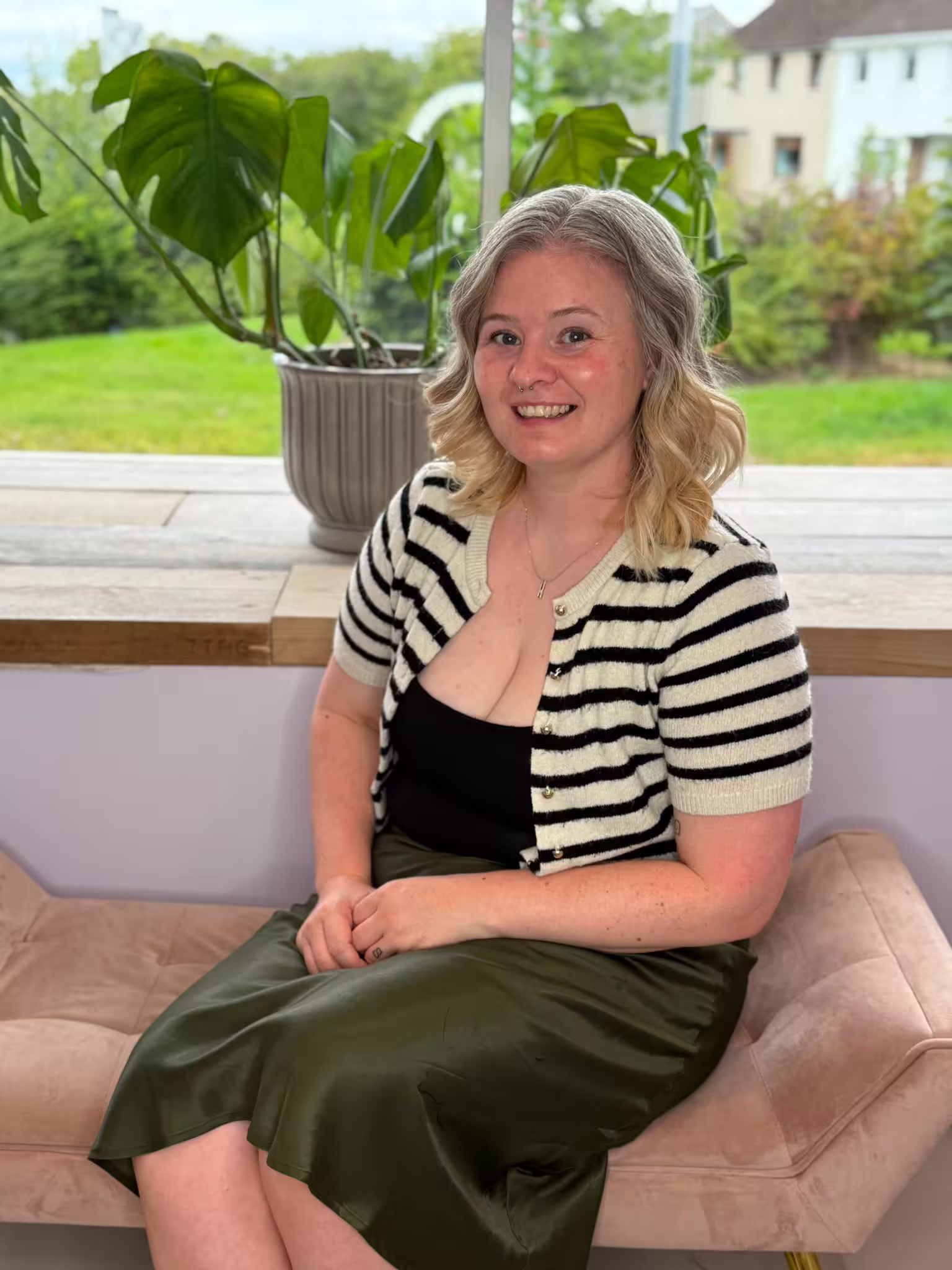 Emma has short, wavy blonde hair, smiles warmly at the camera, wearing a black and white striped knit cardigan and an olive green satin midi skirt