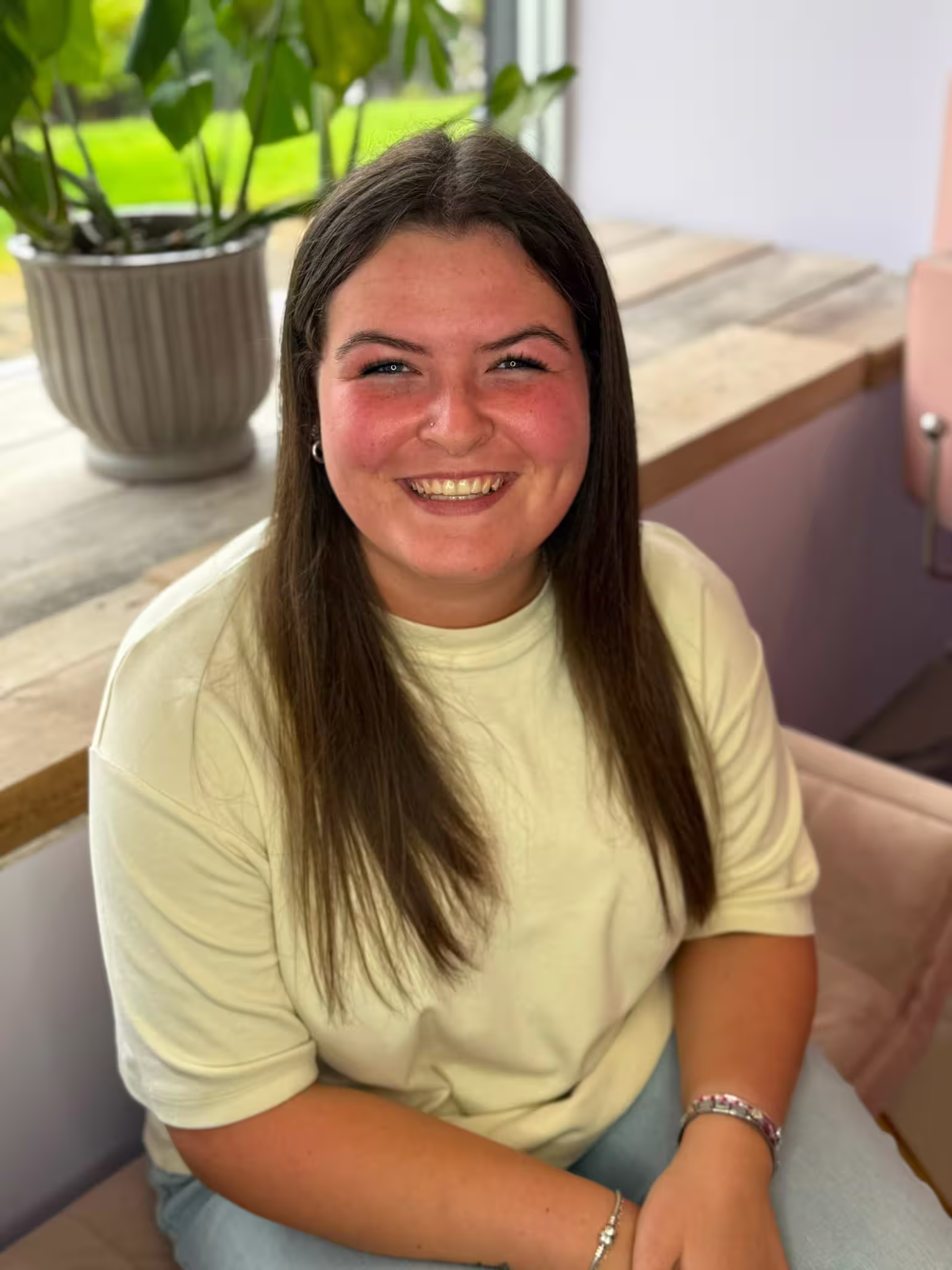 Bobbi has long, straight brown hair smiling at the camera with rosy cheeks, wearing a soft yellow crew-neck t-shirt and jeans