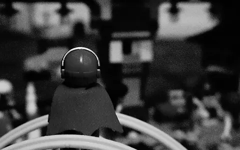 Black and white photo showing the back of a Lego minifigure wearing a helmet and cape, facing a blurred background.