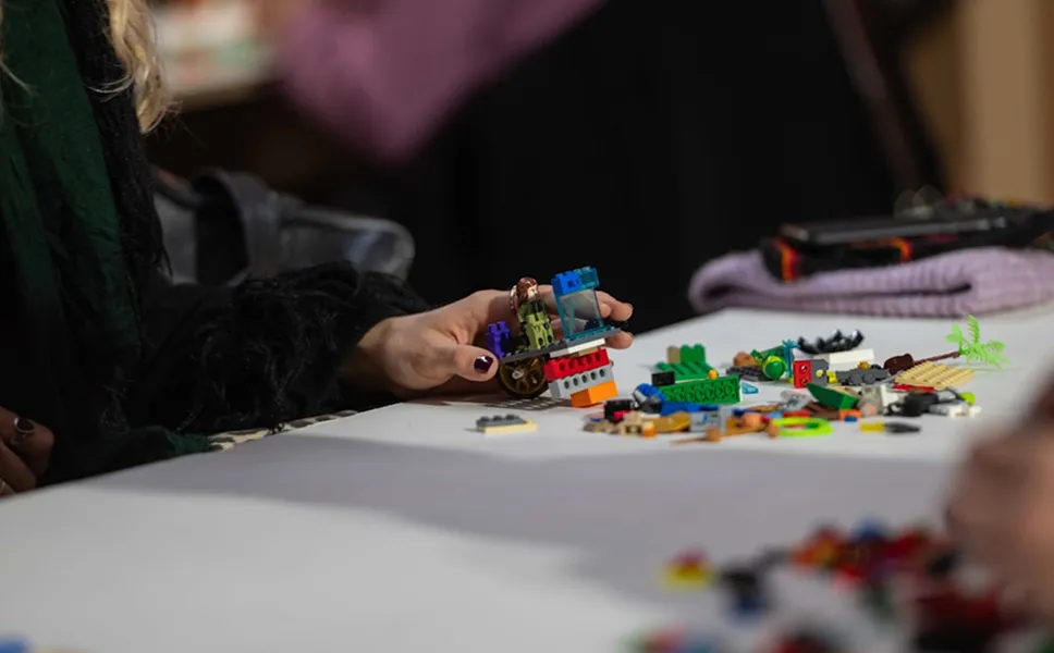 Person assembling colorful LEGO bricks on a white table with scattered LEGO pieces around.