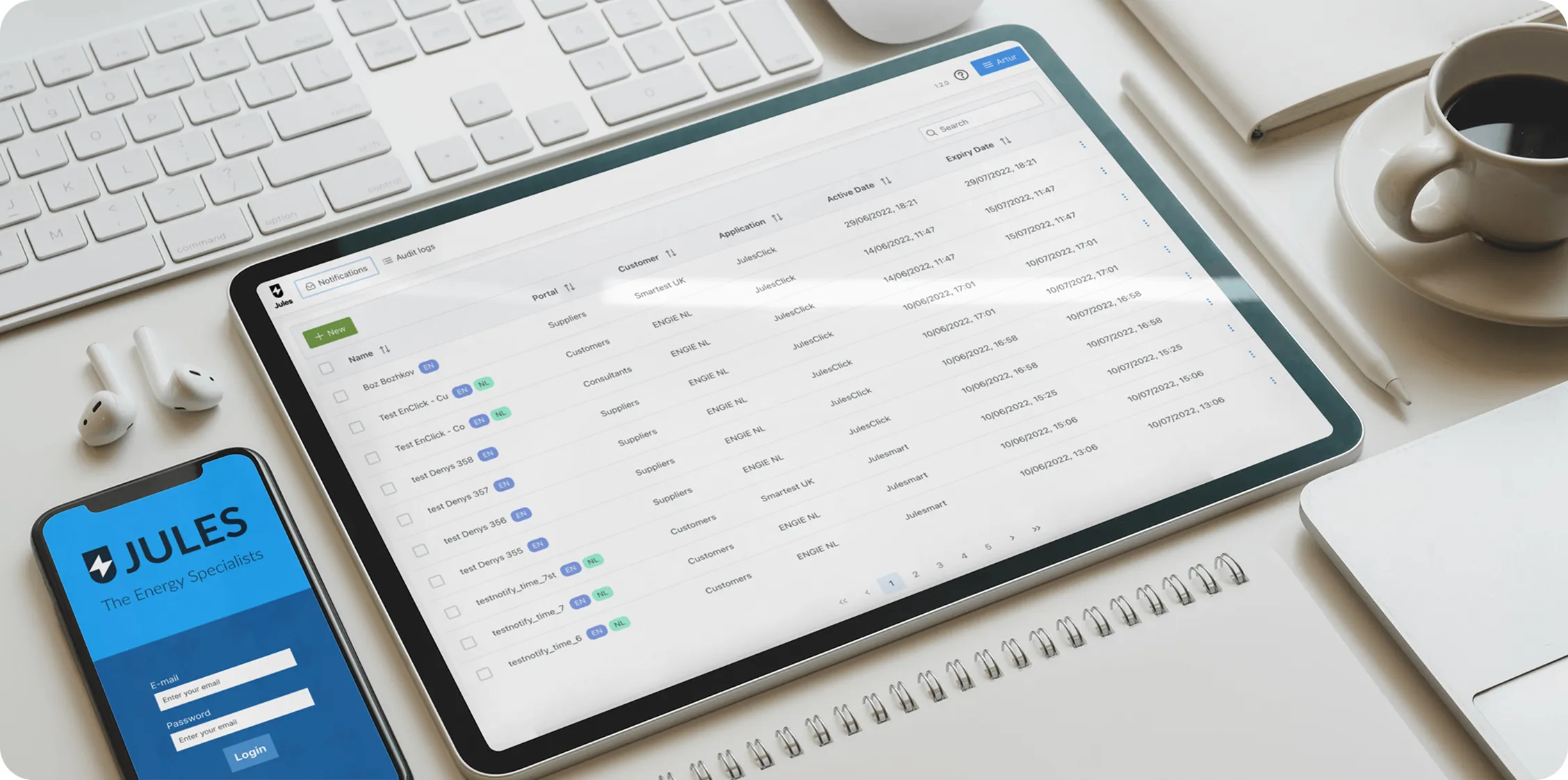 Workspace with a tablet displaying a user interface with data tables, a smartphone showing a login screen for JULES Energy Specialists, wireless earbuds, keyboard, coffee cup, notebook, and pen.
