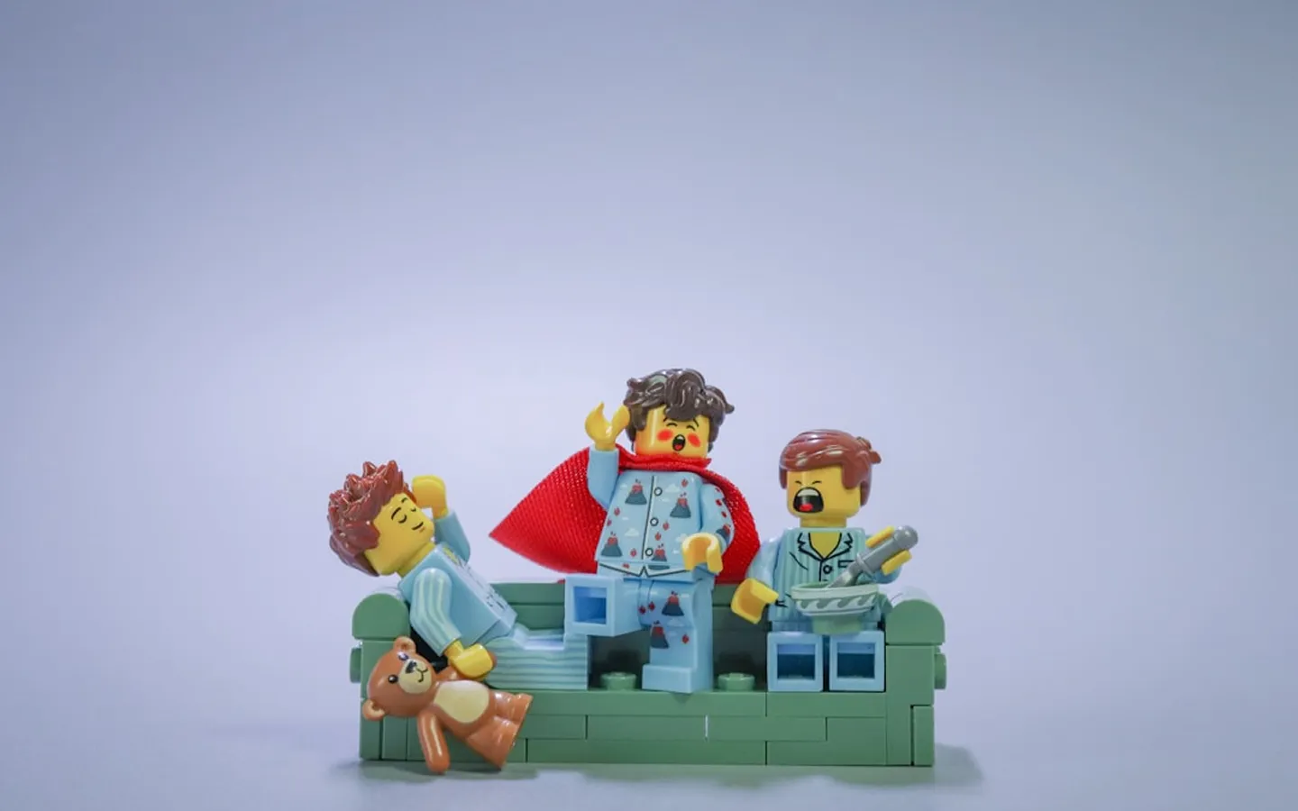 Three LEGO minifigures in pajamas on a green couch; one holds a teddy bear, another wears a red cape, and the third holds a microphone.