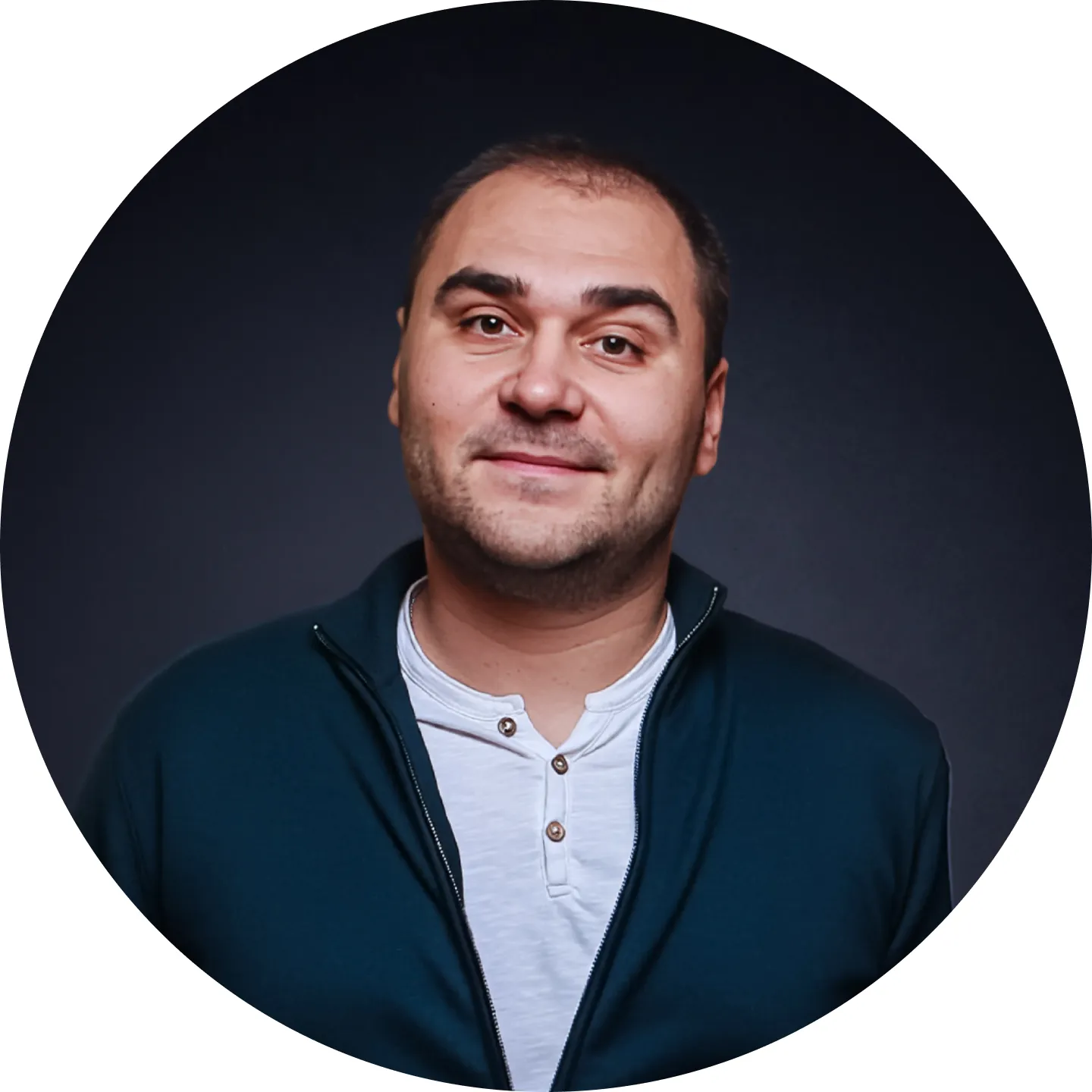 Smiling man with short dark hair wearing a white buttoned shirt and a dark zip-up jacket against a dark background.