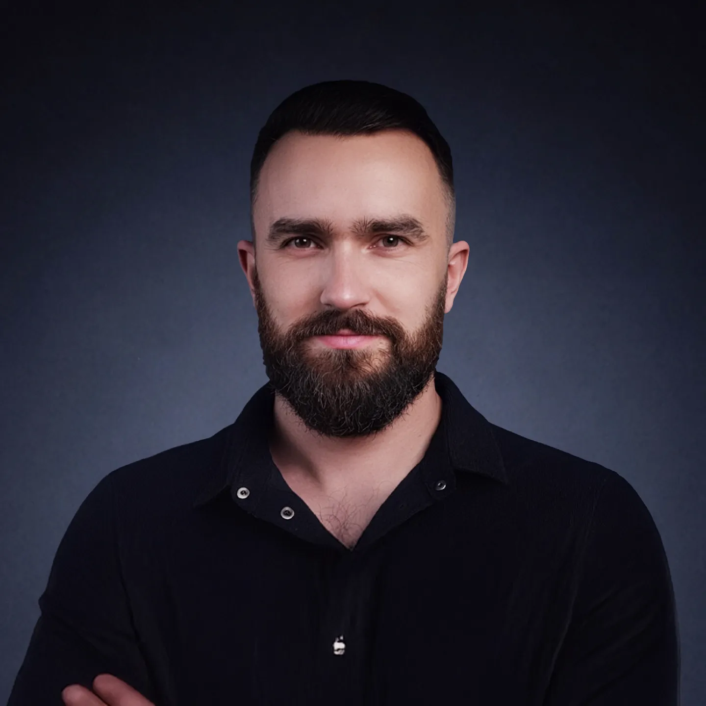 Man with dark hair and beard wearing a black shirt against a dark background.