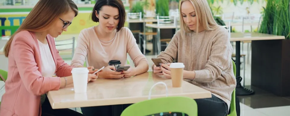 Three women sitting at a cafe table using smartphones with coffee cups in front of them.