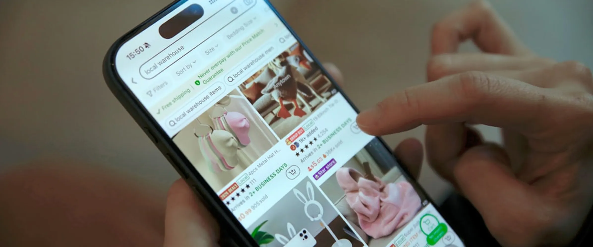 Close-up of a hand browsing local warehouse items on a smartphone screen, showing listings of hats, plush toys, and slippers.