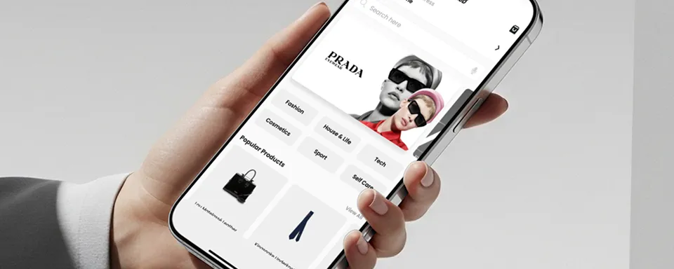 Hand holding a smartphone displaying a Prada online store featuring categories and popular products like a handbag and jeans.